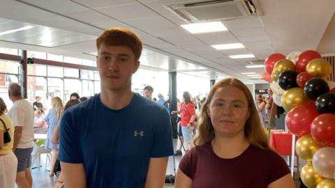 A tall young man with ginger hair and a blue t shirt stands next to his sister, a shorter teenage girl with long auburn hair and a burgundy t-shirt. 