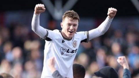 Jude Arthurs on shoulders on fans after promotion, both arms raised as fists abgove his head, smiling wearing Bromley home white kit