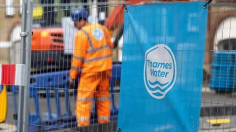 Wire fencing surrounding an area in the street where Thames Water workers are dressed in an orange hi-vis jacket and trousers. There are plastic barriers set up to prevent the public entering that area. On the fencing there is a blue Thames Water sign. 