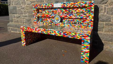 A bench made of thousands of pieces of multi coloured lego bricks. It has rows on the back of the bench which house multiple Lego mini figures. The bench also included a lego version of the blue and white Manchester City football club badge.