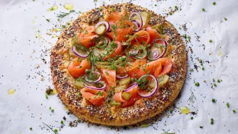 A galette topped with smoked trout, cream cheese and herbs, with seeds scattered all around