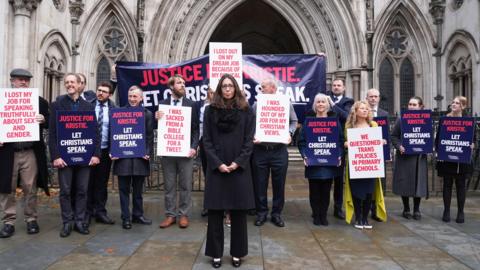 A woman dressed in black stands in front of a group of people outside the Royal Courts of Justice. The group carry placards with messages including "I lost my job for speaking truthfully about sex and gender," "I was hounded out of my job for my Christian views, "I was sacked from a bible college for a tweet" and "we questioned trans policies in primary schools". Others carry signs reading "Justice for Kristie, Let Christians Speak." 