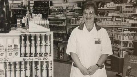 An old, black and white picture of Sadie in her work uniform in the middle of the chemist.