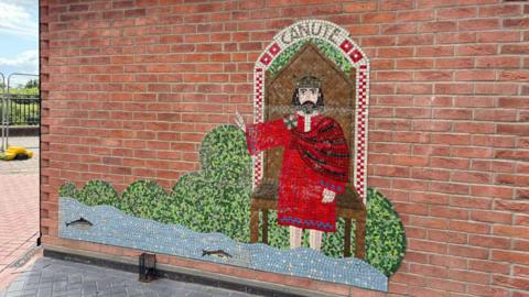 Artwork made of mosaic tiles on a brick wall. It shows a man with a beard, sitting on a brown throne and wearing a red outfit and crown, holding his arm out to motion the tide to stop. Waves and fish are at the forefront and there are trees at the back. The word 'Canute' is written above the throne.
