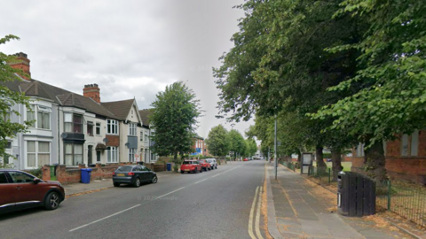 Hainton Avenue in Grimsby. It is a wide road with a pavement and houses on either side. There are a number of park cars in the image, as well as trees and a black bin.
