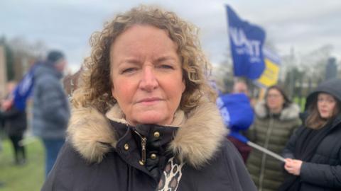 Middle-aged woman with blonde curly hair. She is wearing a fur-hooded parka jacket and stands outside with flags in the background of the picture.