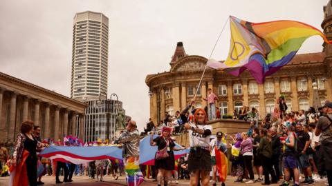 A woman with orange hair waves a rainbow flag in a wide shot Chamberlain Square Birmingham. She is wearing a white t-shirt and black mini skirt. Behind her are crowds of people wearing rainbow accessories. Birmingham's council house can be seen to the left and the Octagon tower behind.