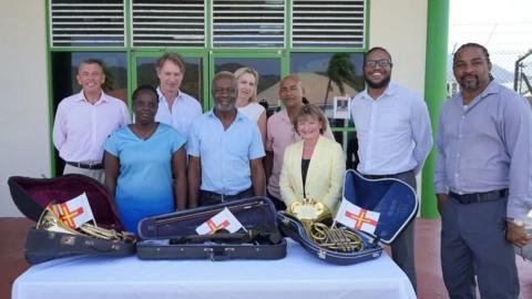 Representatives of Guernsey Music service pose with organisers of a Montserrat music summer school. they are standing as a group, smiling for the camera, standing behind a table with a white table cloth which has three opened musical instrument boxes on it. There are three small Guernsey flags in the boxes.