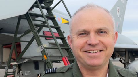 A man wears a dark green RAF pilot uniform and smiles into the camera. He is standing in front of a Typhoon fighter jet. The man has short grey hair and dark blue eyes.