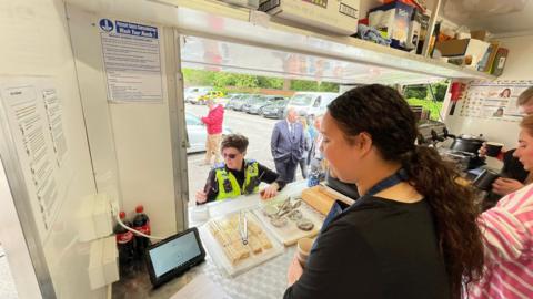A police officer stands at the opening to a food van, looking to scan her card to pay for her purchase. In the foreground, a woman with long dark brown hair can be seen and two others - a man and a woman - can also be seen in the food van.
