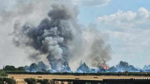 Plumes of grey smoke rising from fields behind a row of trees. Orange flames are visible among the trees.