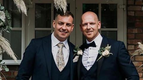 Darren and John George looking into the camera. John ( on the left) is wearing a dark suit, light coloured shirt and patterned tie. His brother is wearing a dark suit, dark bowtie and white shirt. Both men have white flowers on their left lapels and are smiling.