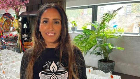 Deepa Parekh is standing in front of rows and rows of paper bags containing food items. She is staring at the camera and is wearing a black hooded jumper with a candle graphic on the front. 