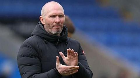 Shrewsbury Town head coach Michael Appleton is wearing his black puffer coat and clapping the crowd after a game
