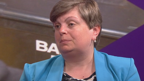 A woman wearing a black and white patterned top and a light blue suit jacket is sitting on a red sofa. She is looking to the left of the shot.