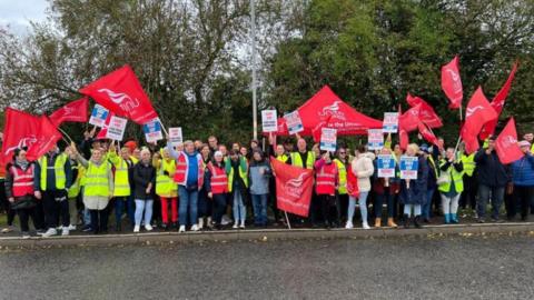 A crowd of striking workers outside the Spalding factory. They are mainly wearing hi-vis jackets and have red Unite the Union flags and placards.