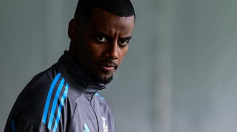 Alexander Isak is seen from chest up in a black and blue zip-up Newcastle United training top, dipping his head and looking directly at the camera with the background blurred out