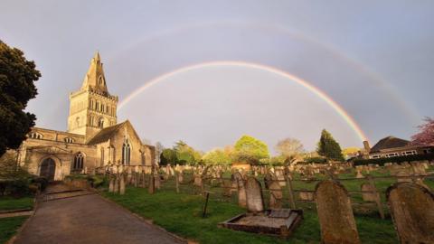 A double rainbow in the grey sky over the cemetery graves and church behind the cemetery.