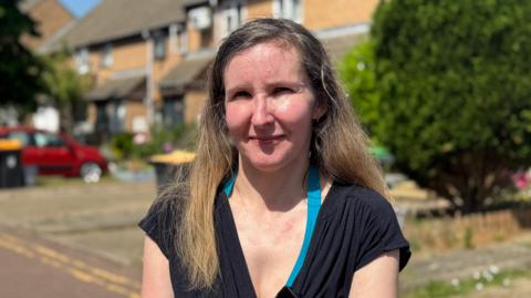 Julia is standing in a street of terraced homes. She has long hair and is wearing a black short-sleeved dress and is squinting slightly in the strong sunshine. 