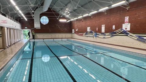 A large swimming pool inside a brick building with a pointed metal roof and changing rooms to the left hand side