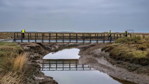 A general view of Stiffkey bridge - a footbridge crossing a marshland creek. There are three workers on the bridge.