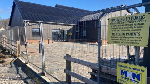 Site of a community centre construction project. The building is fenced off. There is a warning sign telling people not to enter the site. The building is dark coloured with solar panels on the roof.