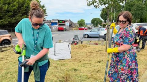 A woman in a turquoise T-shirt and black sunglasses trims grass, while another woman in a flowery dress rakes the grass.