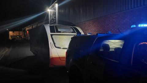 A police car parked by the caravan at Banbury train station. It is night time. The police car light is flashing blue. 