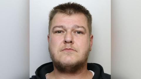A man in a black hoodie with a earring in his left ear looks at the camera as part of a police mug shot