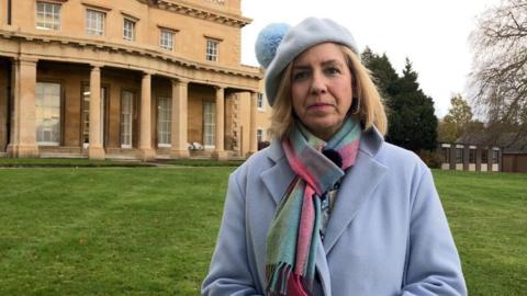 Dame Andrea Jenkyns standing on a grassed area in front of a grand building. She is wearing a blue winter jacket and matching bobble hat, along with a multi-coloured scarf.