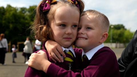 Young boy hugs his twin sister, they are wearing a red and yellow school uniform