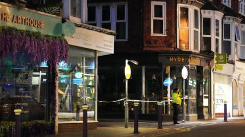 A police woman stands outside a row of shops in an area that has been closed off to the public. It is night time.