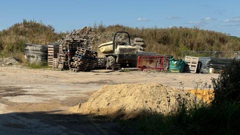 The site where allotments are meant to be built photographed on a sunny day. There is a pile of dirt, construction materials and a vehicle parked there. A bit of greenery could be seen in the background.