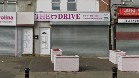 A street-view image of the charity's former base on Finch Lane. It is a shop front with metal shop-front barriers and a sign above the door reads "The Drive".