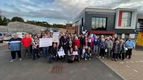 A group of about 70 people holding banners are stood outside Loggerheads fire station. 