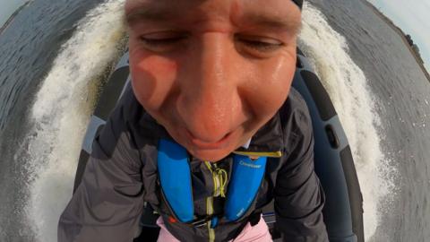 Wide angle close up of a man's smiling face, sitting in a speeding boat with wake and whitewater around the boat.
