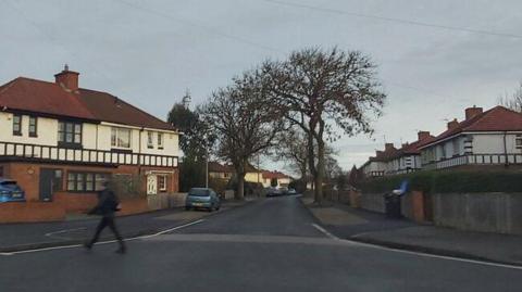 A photo of Musgrave Gardens, taken from Sunderland Road. Musgrave Gardens is a narrow street lined by two storey semi-detached houses with white top facades. There are several trees and parked cars. One person is crossing the street.