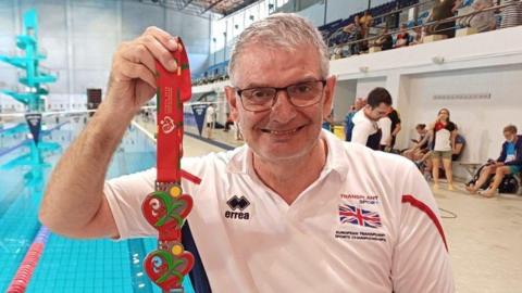 Rob Hodgkiss holding up his medals in front of a swimming pool