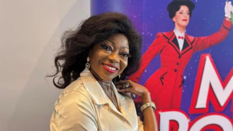 Patti Boulaye at the Birmingham Hippodrome. In the head and shoulders shot she is wearing a cream coloured shirt and is smiling.  Behind her is part of a red and purple advertising board for Mary Poppins