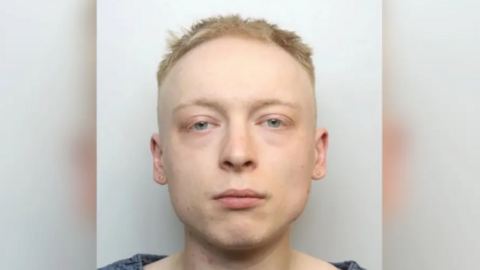 A mugshot of Miller, who has a vacant expression and is looking at the camera. He has short blonde hair and is wearing a blue top.