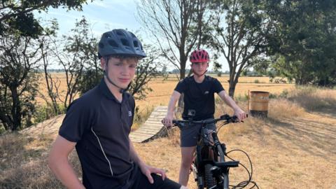 Two teenagers stand side by side on bikes wearing cycle helmets