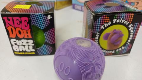 A purple toy in the shape of a tennis ball is pictured in front of brightly coloured boxes.