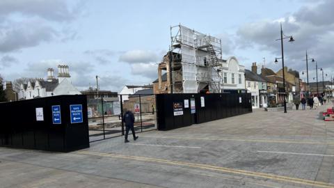 One section of a building is clad in scaffolding and white sheeting about 30 feet (9m) high. A black fence surrounds the partly demolished structure, some people are walking along the wide pavements, with ornate black lampposts visible too.
