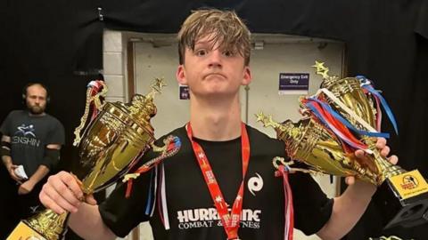 A teenage boy stands wearing a black t-shirt and holding three gold trophies and with a medal around his neck.