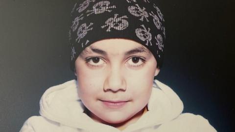 A young boy with a black hat and white hoodie looks at the camera