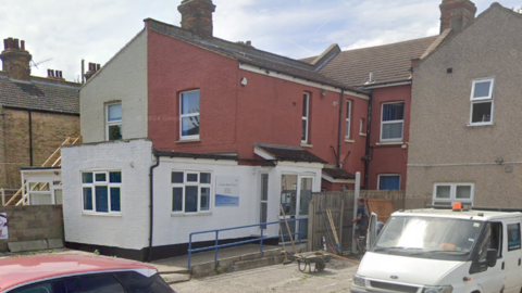 A red and white terraced building is seen in the middle of the picture. The bottom half of the building is white, with a rail leading up to the door. The top half is red, and shows multiple windows. 