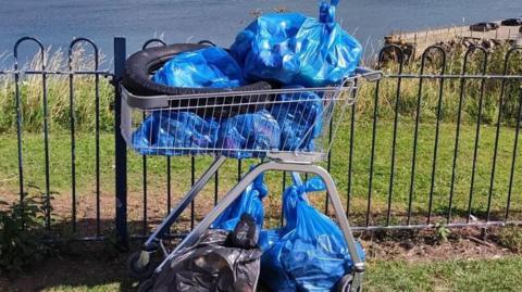 A tyre and blue bags of rubbish in a shopping trolley beside a metal fence overlooking a beach. The blue sea is visible in the background. 