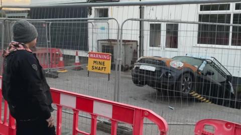 A man looks through fencing at his car. The rear of the Mini has fallen into a sinkhole. On the fencing is a sign which reads "Warning mineshaft".