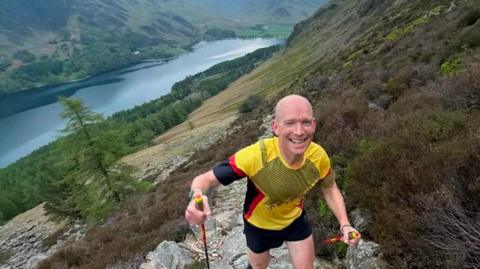 Runner Kim Collison wears a yellow top and black shorts and he ascends up a path. He is smiling as he ascends the mountain path in the Lake District. There is a large lake behind him.