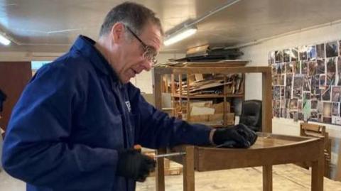 A man in blue overalls holding a screwdriver to the seat of a wooden chair. He has black greying hair and is wearing glasses. He is standing in what appears to be a store room, the chair placed on a large wooden table, with assorted items of furniture and off-cuts on shelves around him.